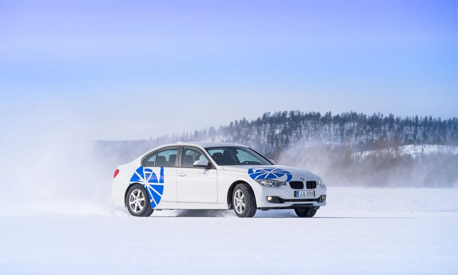 A white sedan with blue geometric decals driving on snow-covered landscape with a snowy forest and blue sky in the background.