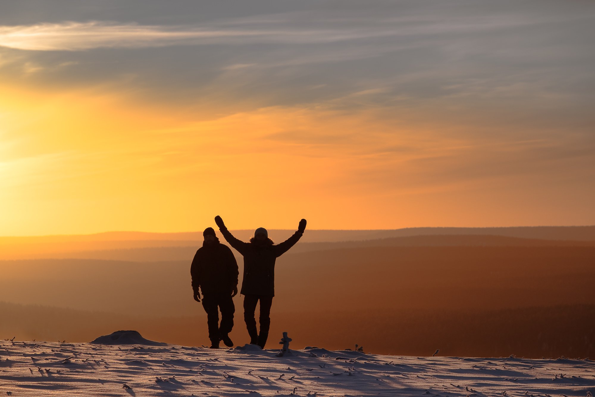 Two people walking on snow-covered ground during sunset, with one person raising arms in celebration against a colorful sky.
