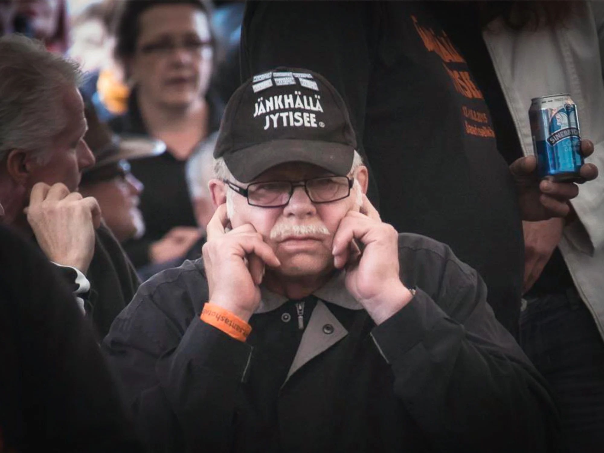 An older man with glasses and a white mustache holding his ears with his fingers at a crowded event.
