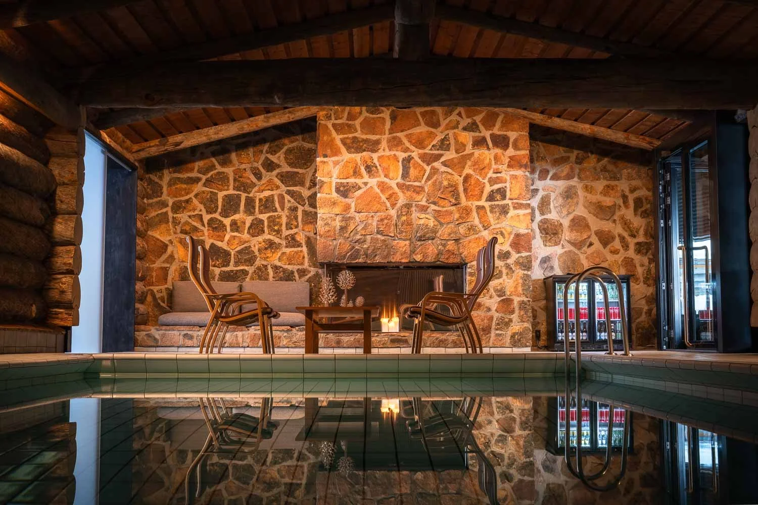 Indoor pool area with stone fireplace, chairs, small table, and reflective water surface.