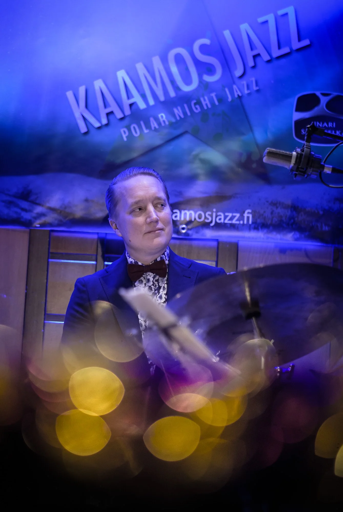 A man dressed in a suit with a patterned shirt and bow tie playing the drums at a jazz event called Kaoamos Jazz, with colorful bokeh lights and a microphone in the background.