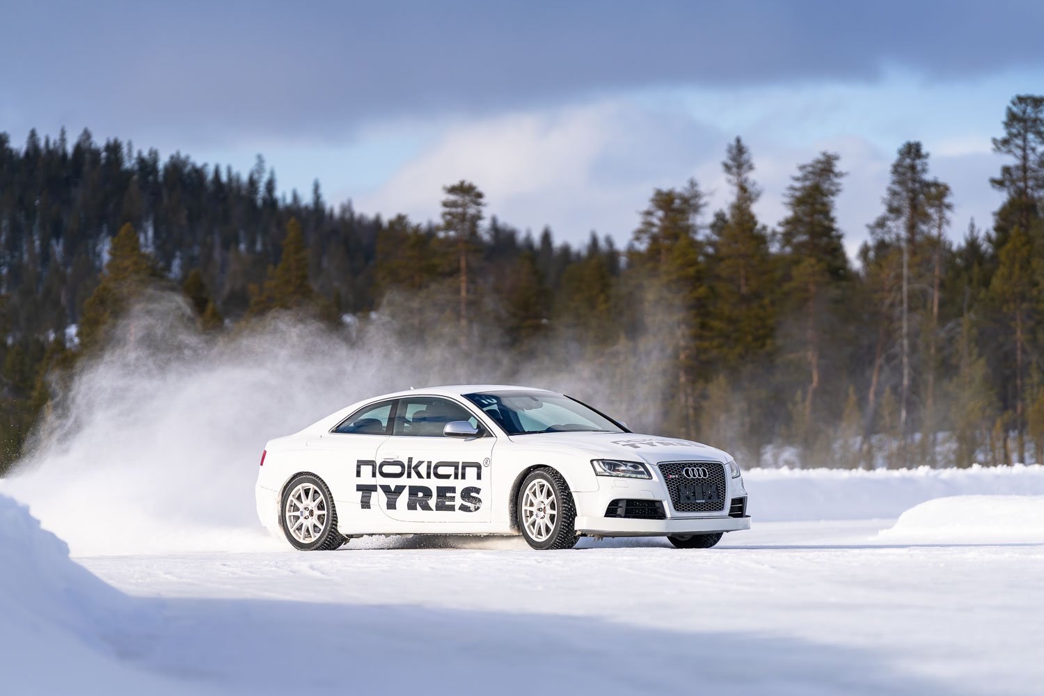 White car with Nokian Tyres logo driving on snow with a spray of snow behind it, coniferous trees in the background, overcast sky.