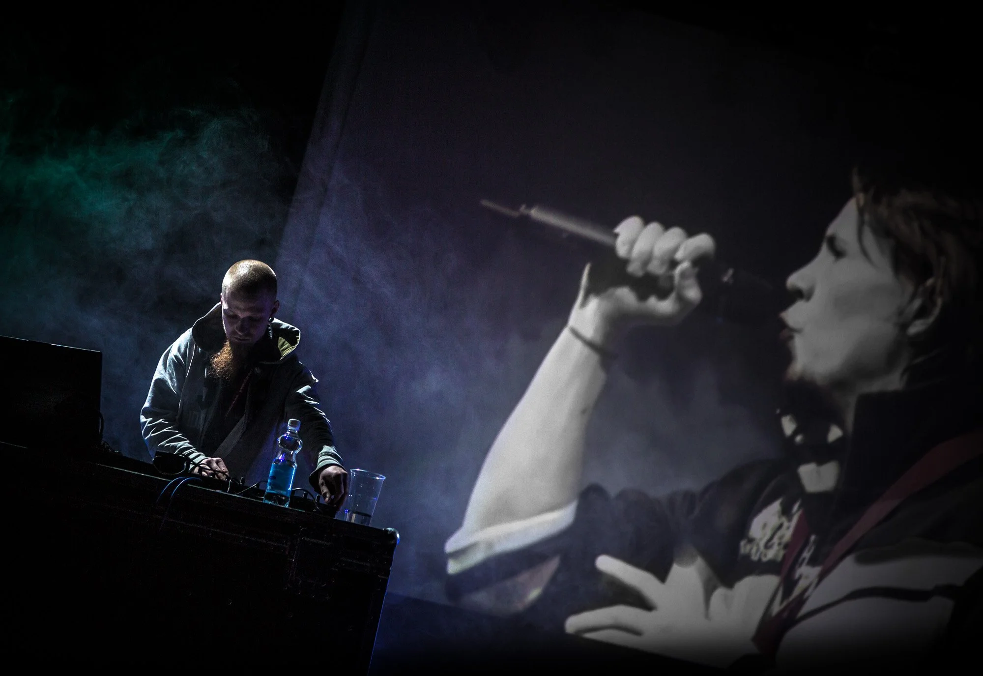 A performance with a DJ on the left, and a large black and white projection of a woman singing into a microphone on the right.