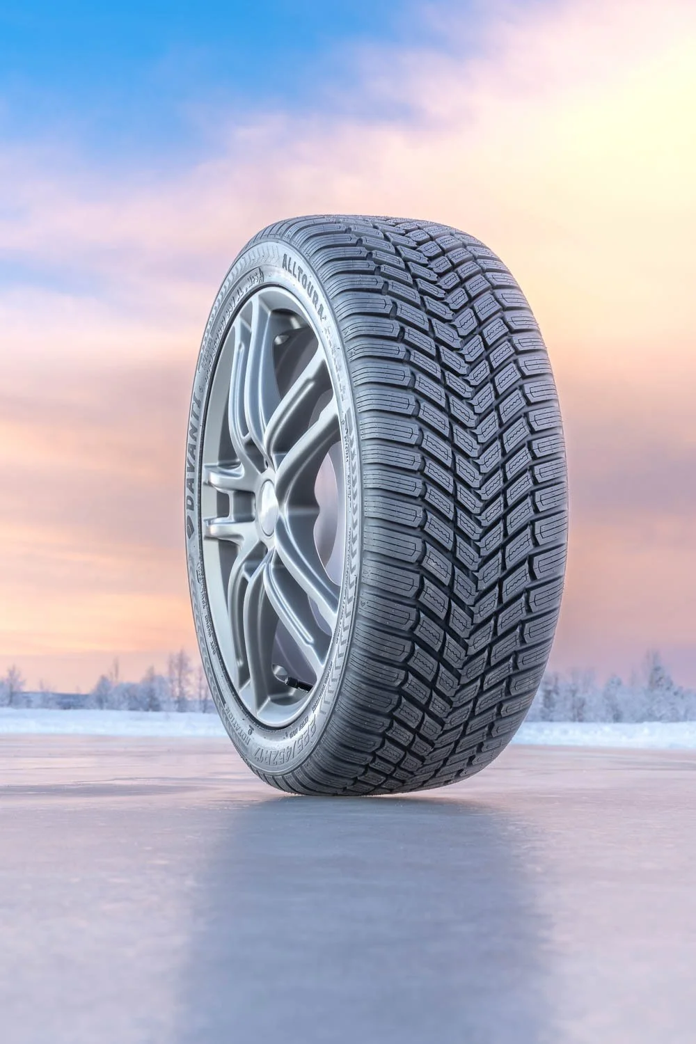 A car tire with a detailed tread pattern, mounted on a silver wheel, set against a winter landscape with snow and a colorful sky.