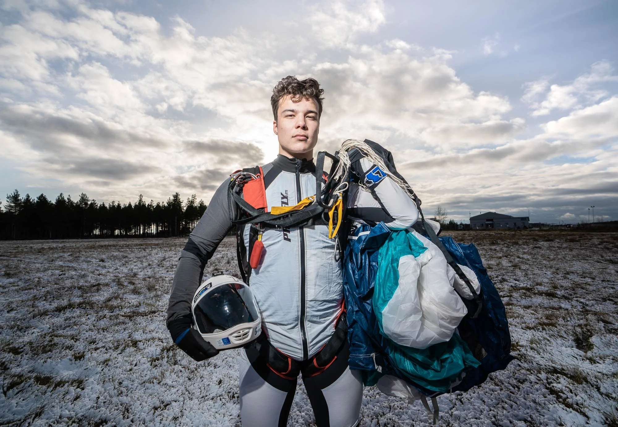 Young man in outdoor gear standing in snow-covered field, holding a helmet and a large backpack with survival gear, with cloudy sky and distant trees in the background.