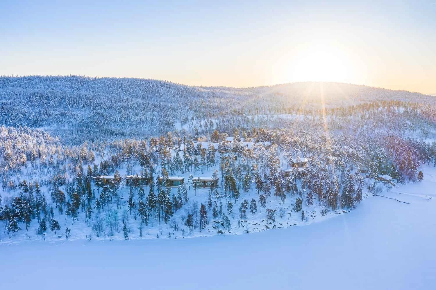 Snow-covered forested mountains with a few houses and the sun rising in the background.