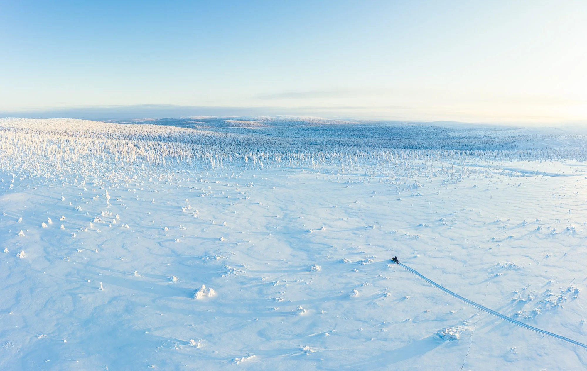 A vast snowy landscape with a lone skier creating a trail in the snow, surrounded by snow-covered trees and distant hills under a clear blue sky.