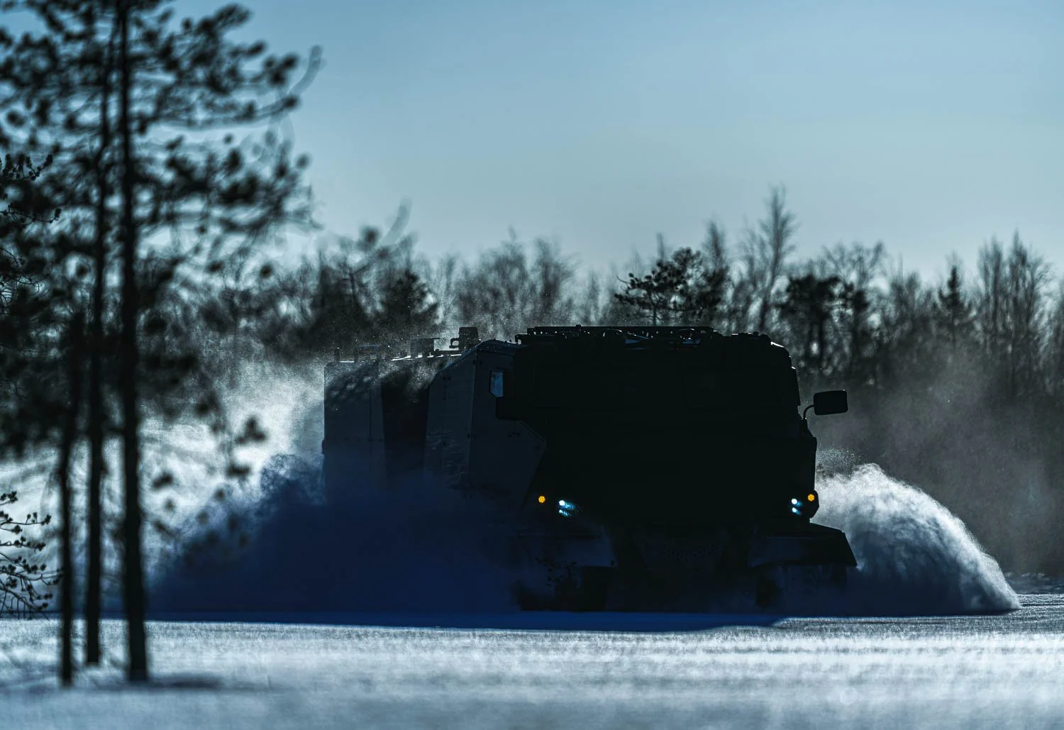 A military armored vehicle driving through snow, creating a spray of snow behind it, with trees and a cloudy sky in the background.