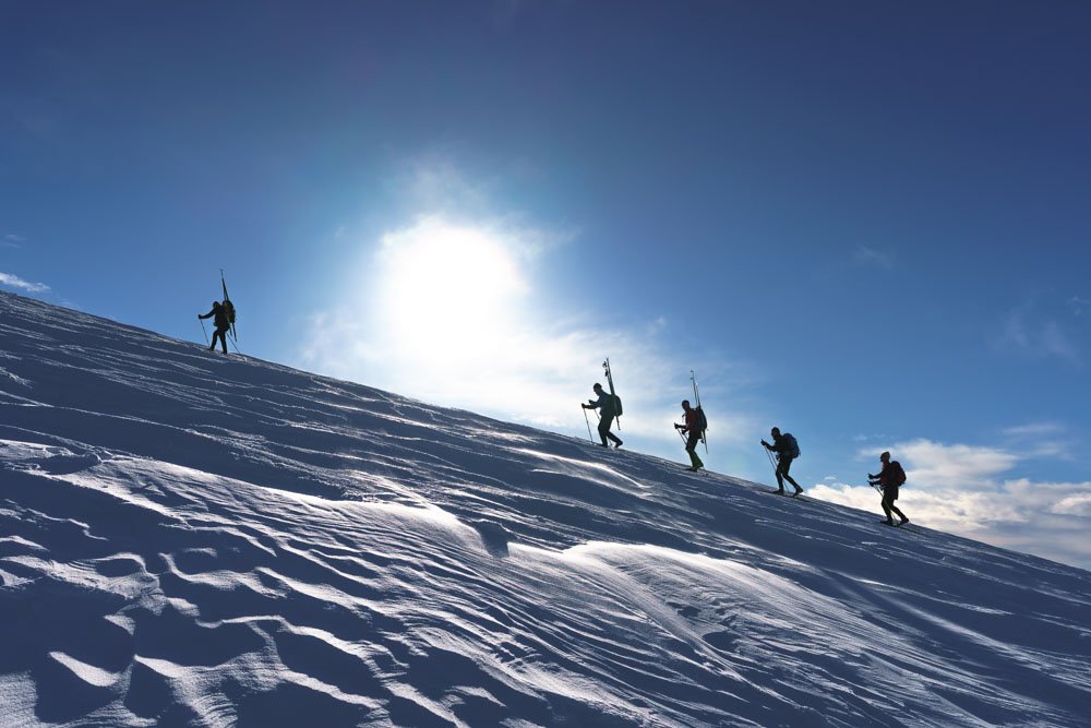 Group of five skiers ascending a snowy mountain slope with the sun behind them and a clear sky above.