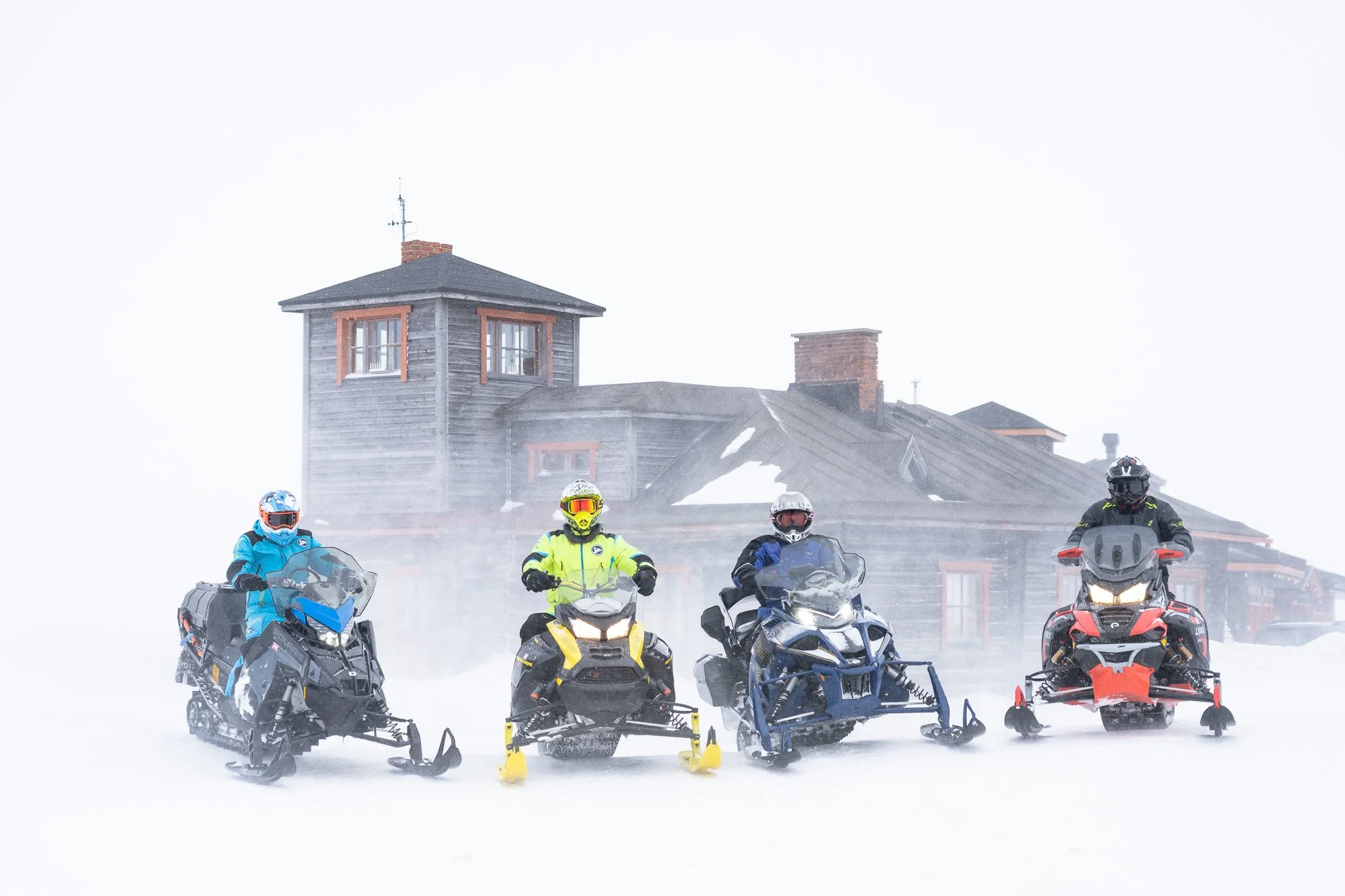 Four people in winter gear on snowmobiles in front of an old wooden house on a snowy day.