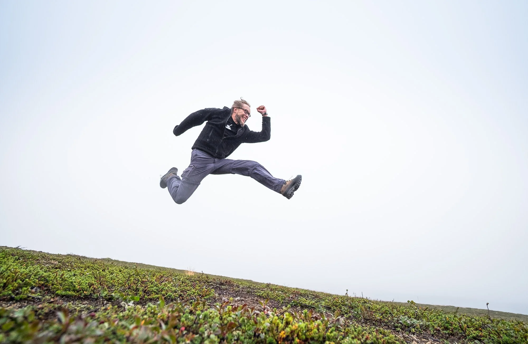 A man in outdoor clothing is mid-air jumping on a grassy hill with a cloudy sky background.