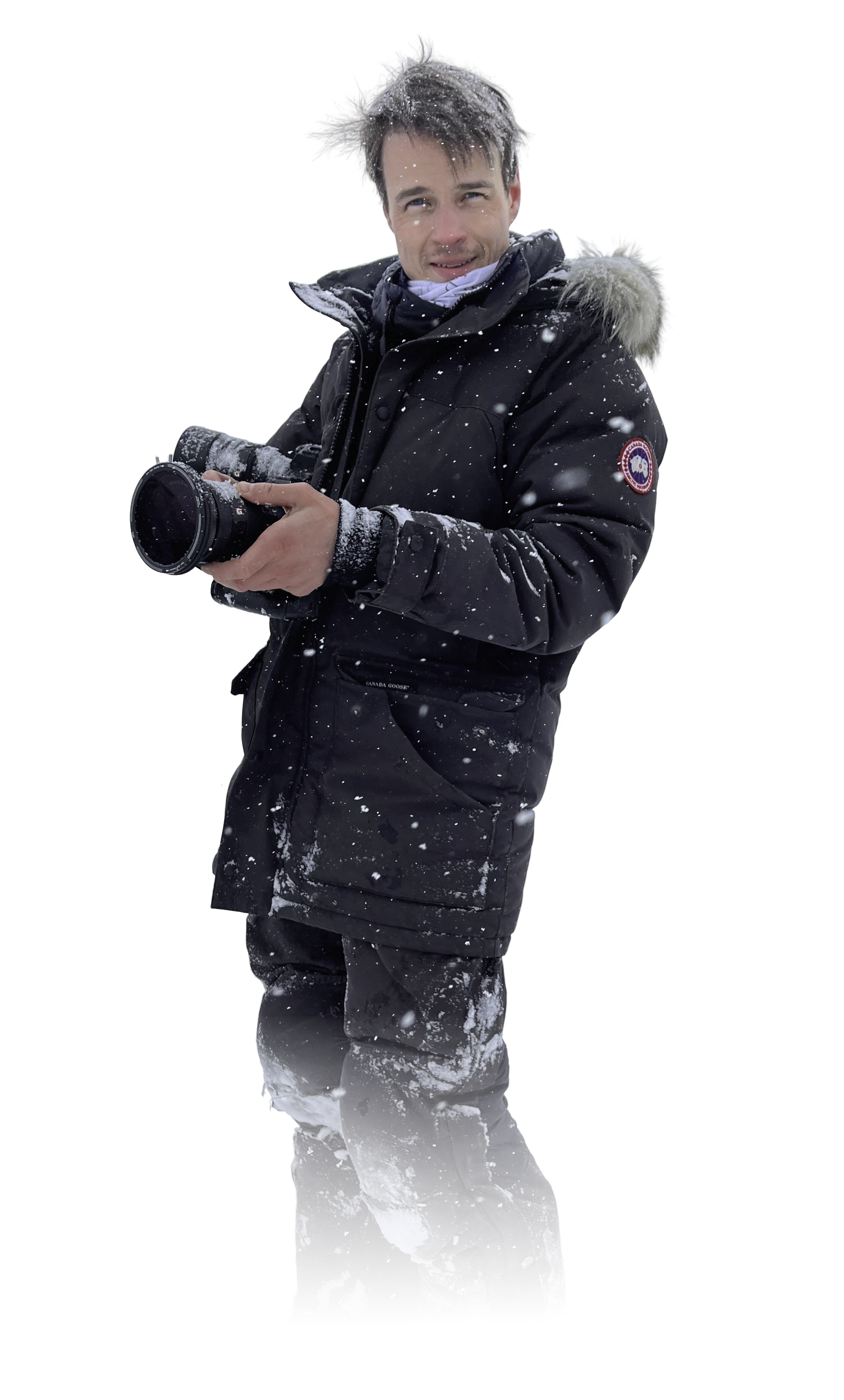 A man standing outdoors in snowy weather, wearing a black winter jacket with a Canada Goose logo, holding a professional camera, with snow on his hair and clothing.