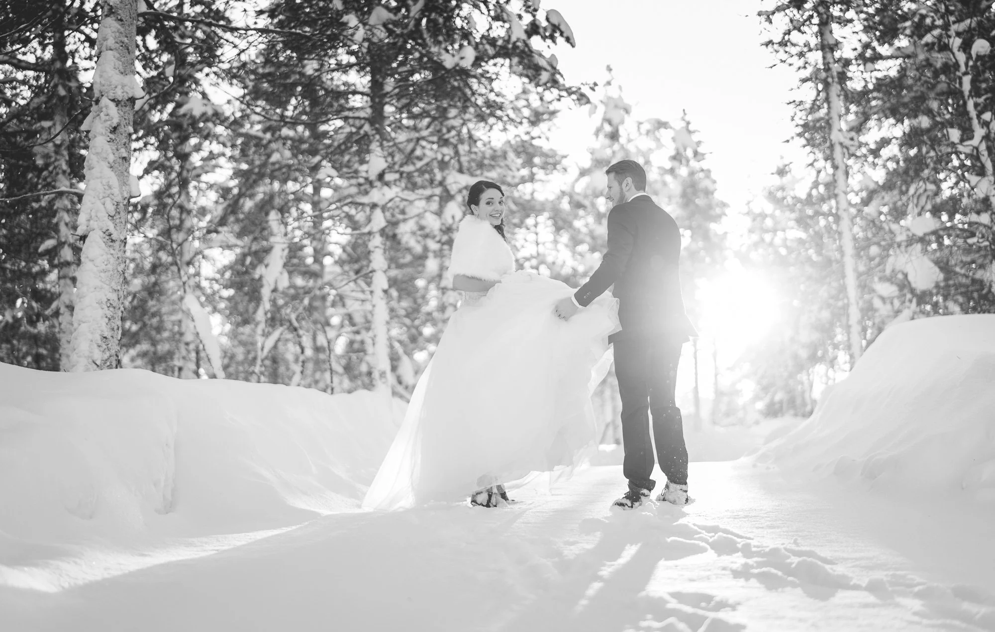 A bride and groom holding hands in a snowy forest, with sunlight shining through the trees in the background.
