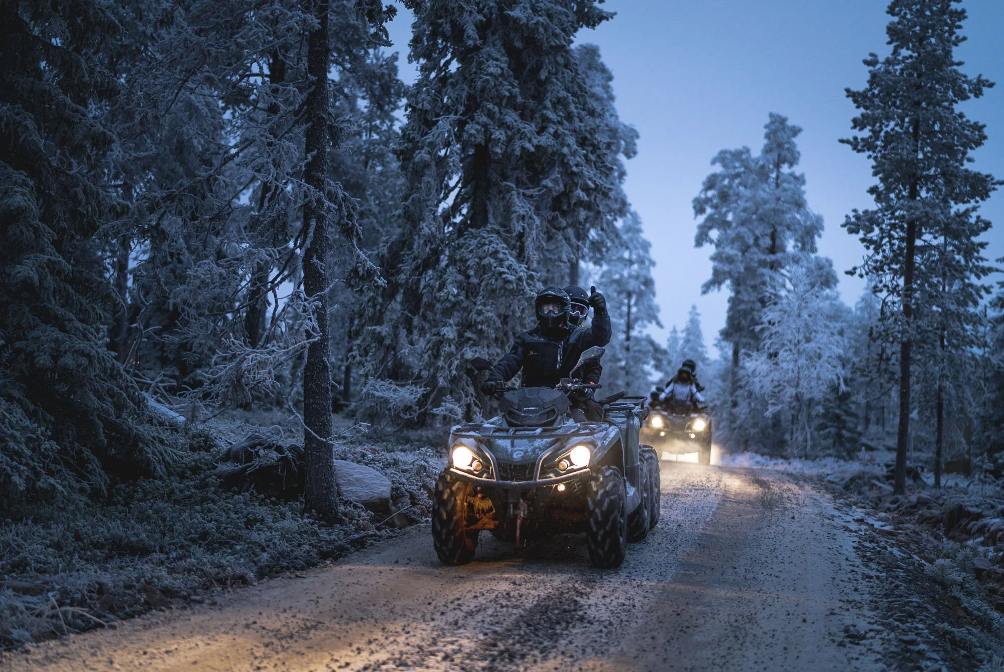 A group of people riding all-terrain vehicles through a snowy forest path during twilight, with the rider in front wearing a helmet and gloves, waving.