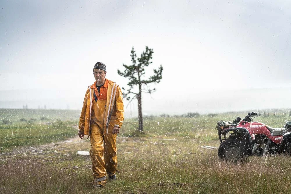 A man wearing a yellow rain gear walking in a grassy field during rain, with a tree on his left and a red all-terrain vehicle on his right.