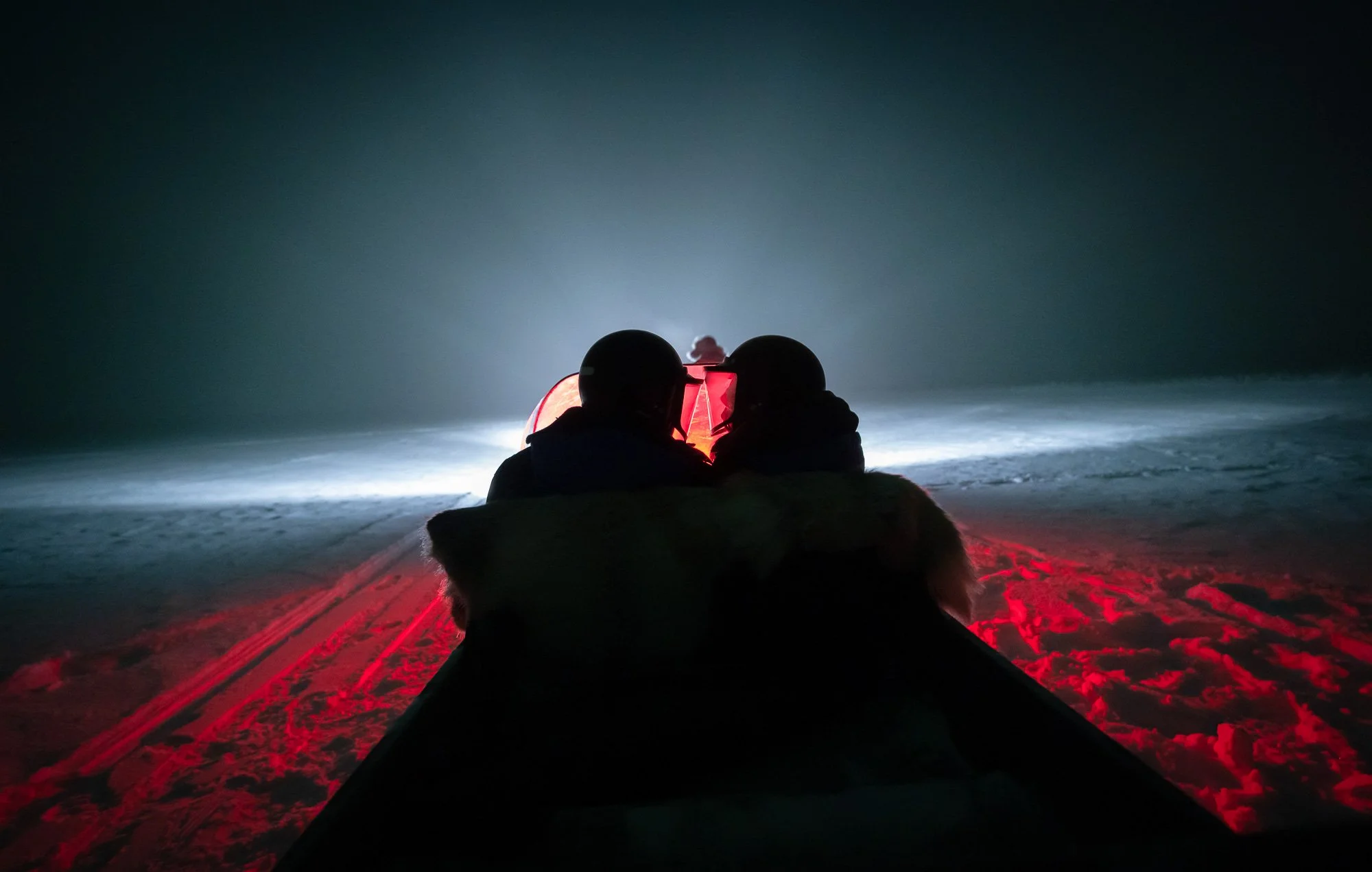 Two people wearing helmets on a snow-covered terrain at night, illuminated by the red and white lights of a sled or vehicle, with a dark, foggy background.