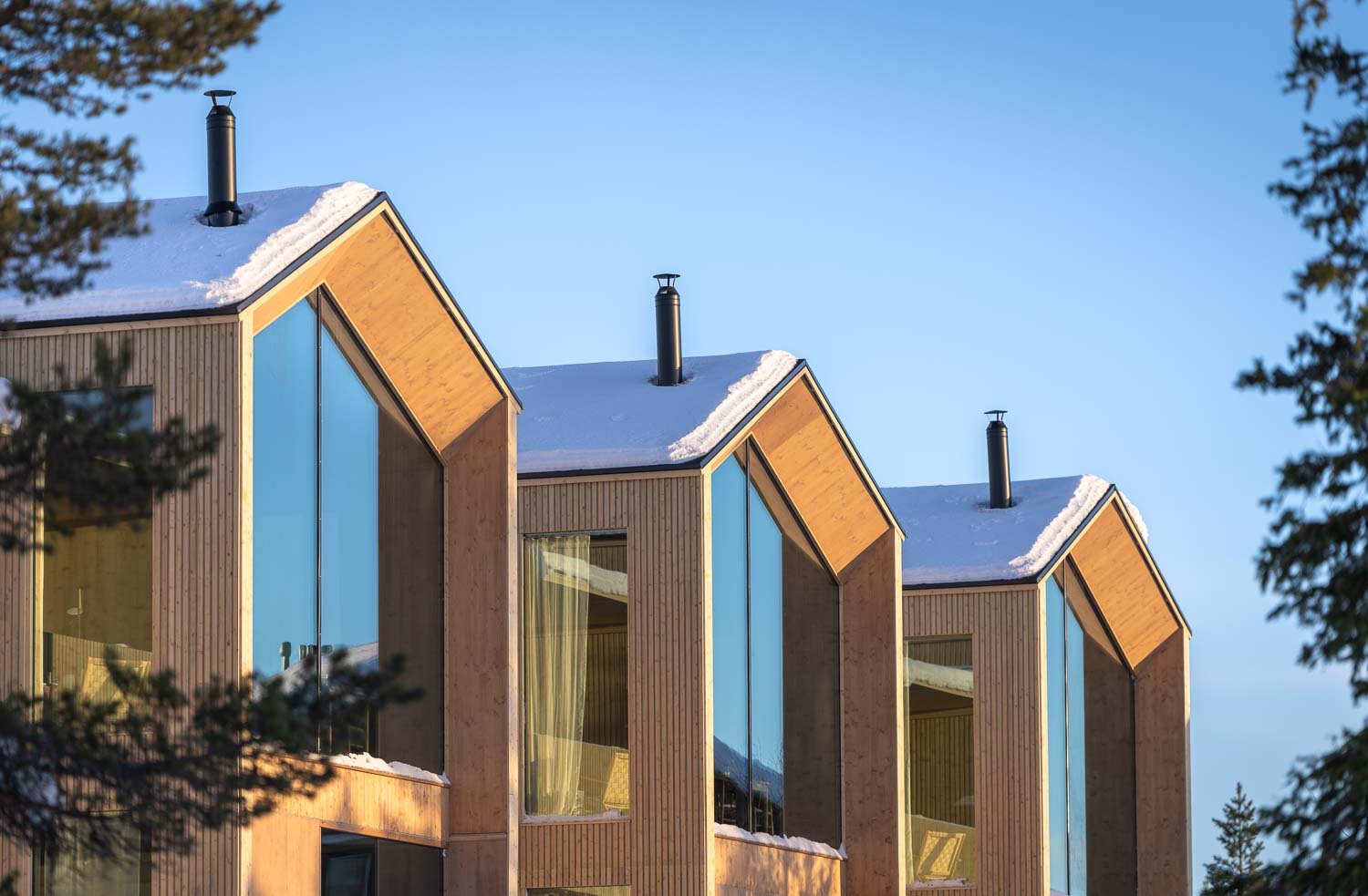 Modern residential buildings with large glass windows and sloped roofs covered in snow, with trees in the foreground and clear blue sky in the background.