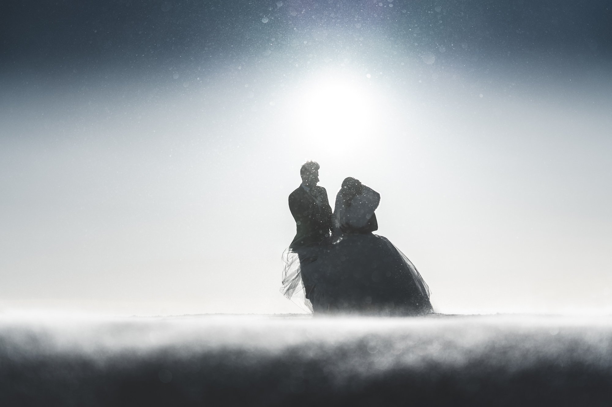 Silhouettes of a man and woman in wedding attire standing close together in a snowy landscape with a bright sun or moon in the sky, creating a dramatic backlit scene.