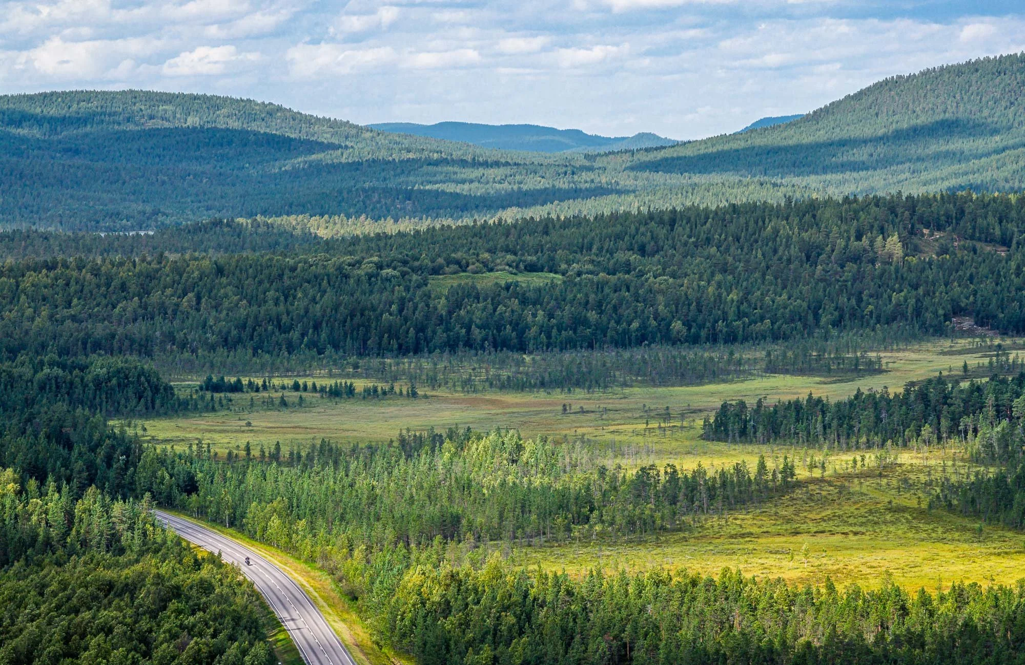 Lush green forests and rolling hills with a highway cutting through the landscape under a partly cloudy sky.