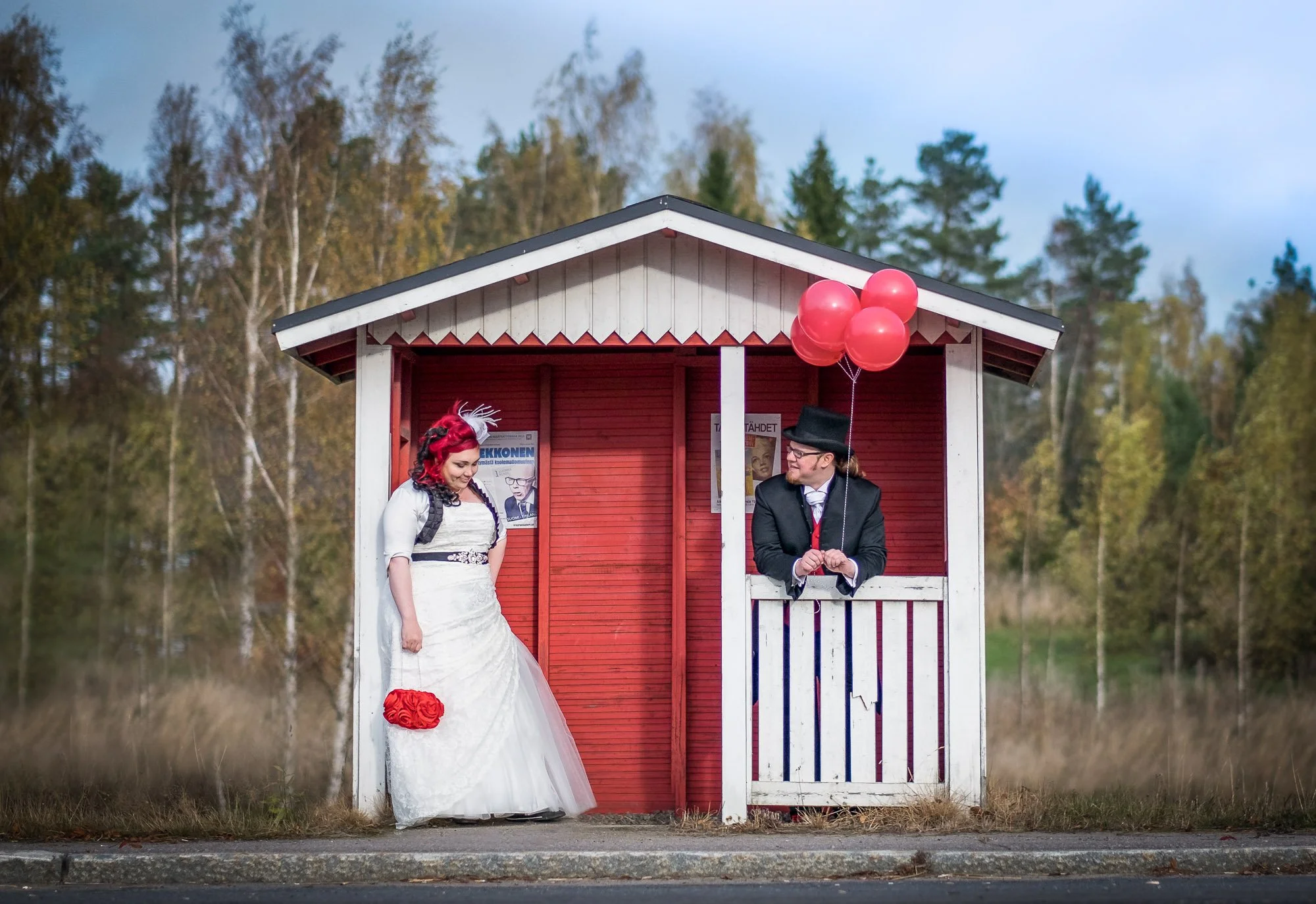 A bride and groom in vintage attire standing outside a small red and white wooden bus stop shelter with posters, holding red balloons.