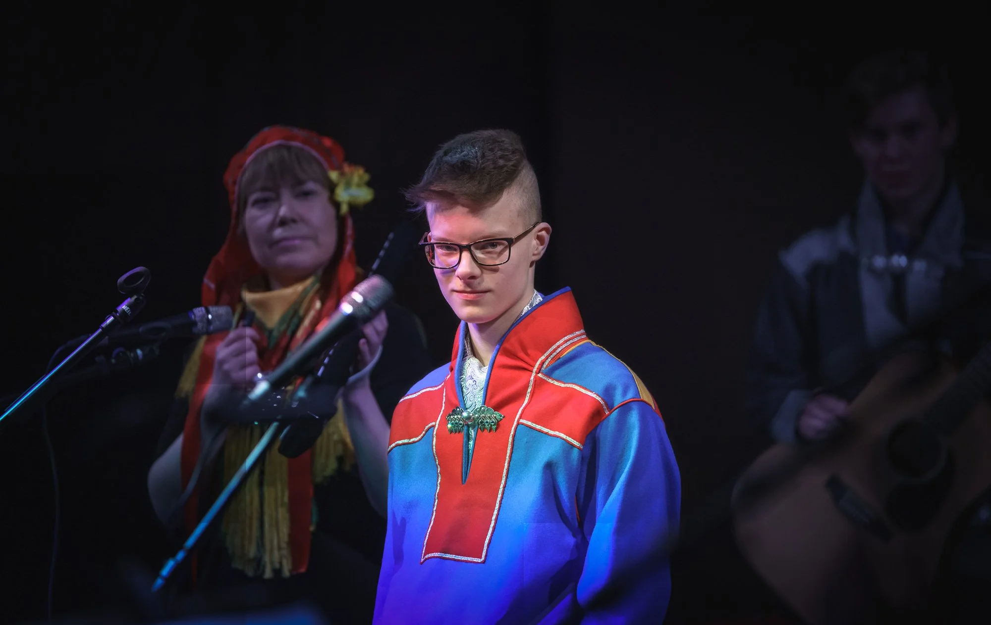A young man wearing glasses and a colorful traditional outfit stands on stage, with a serious expression. In the background, a woman in traditional attire holds a microphone, and another person with a guitar is partially visible.
