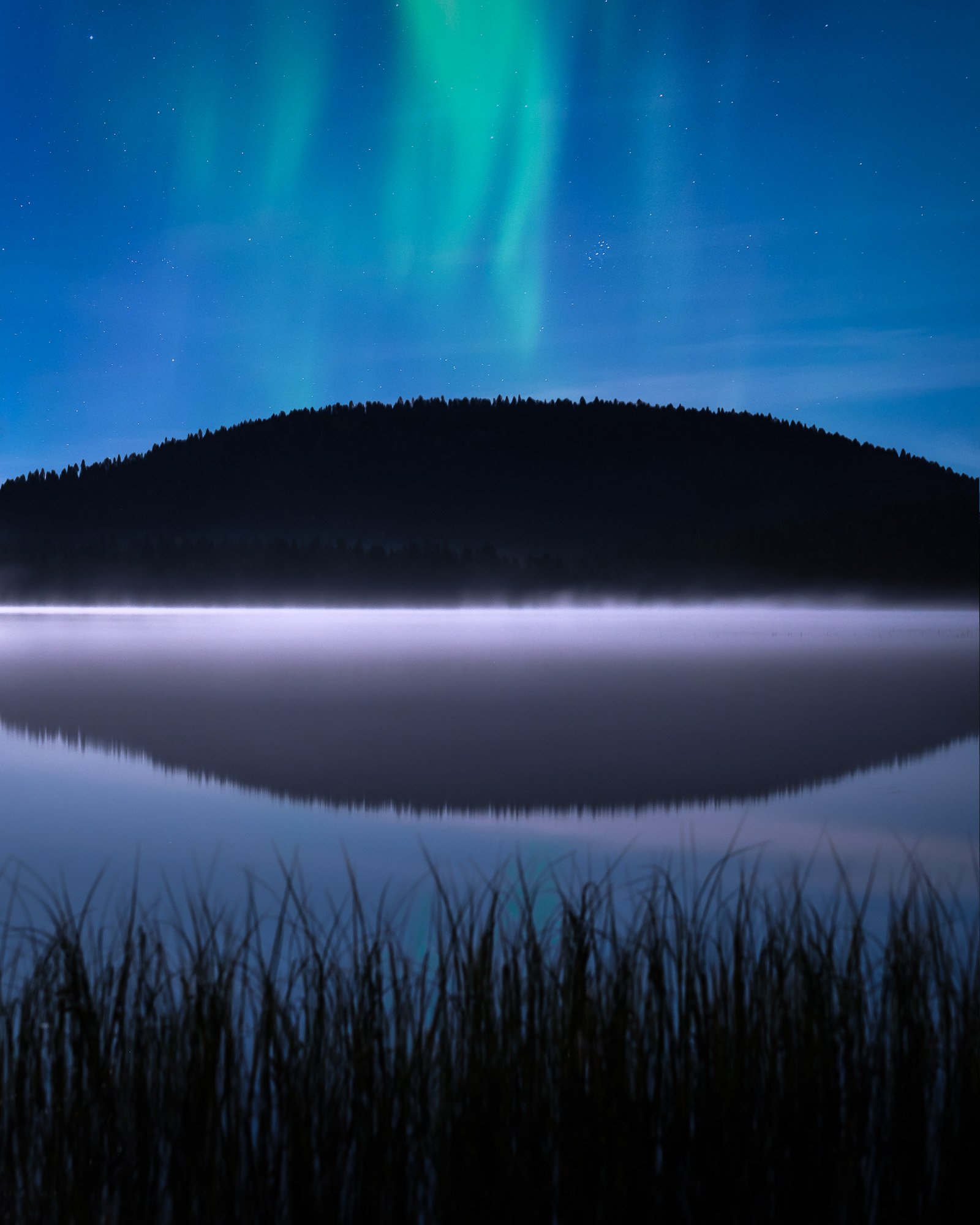 Northern lights in the sky above a lake with a distant tree-covered hill and its reflection in the water, with grass in the foreground.