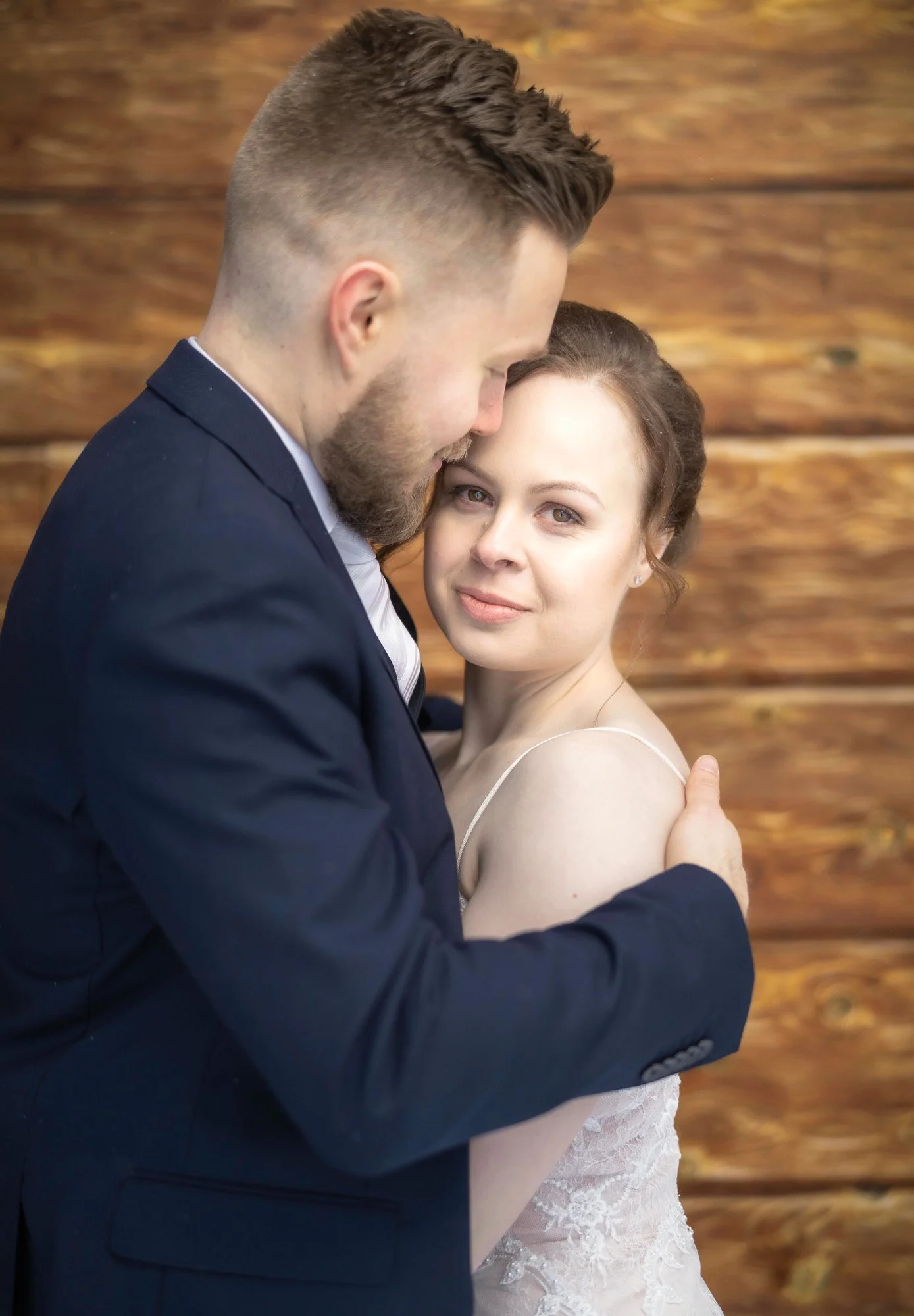 A couple dressed in formal attire embracing each other, with a wooden background.