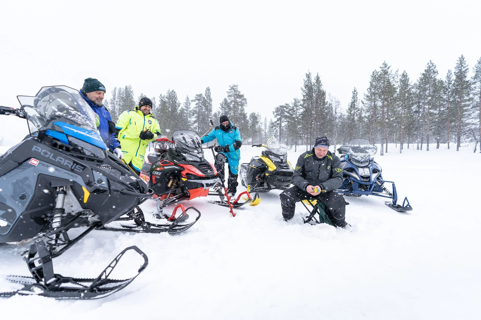 Group of six people with snowmobiles in a snowy, wooded landscape.