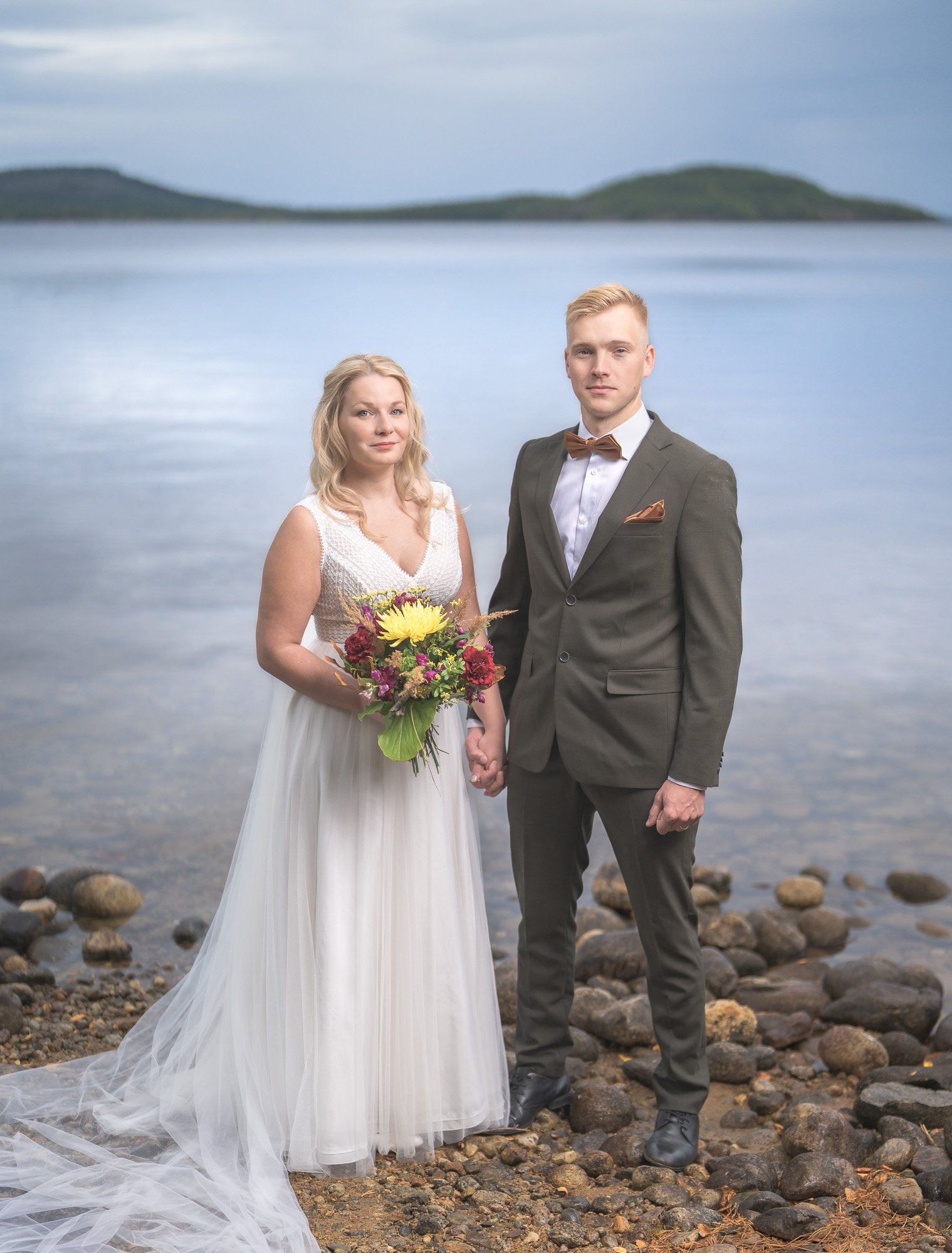 A bride and groom holding hands by a rocky lakeshore with a blurred island and water in the background.