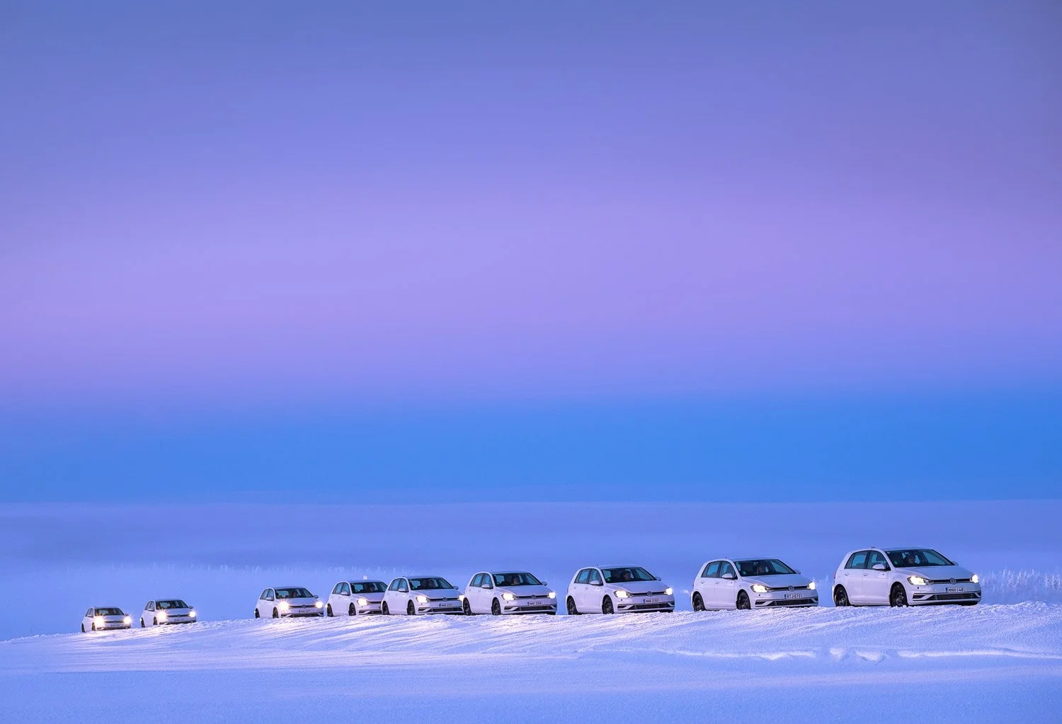 A lineup of eight white cars parked on snow under a pastel purple and blue sky.