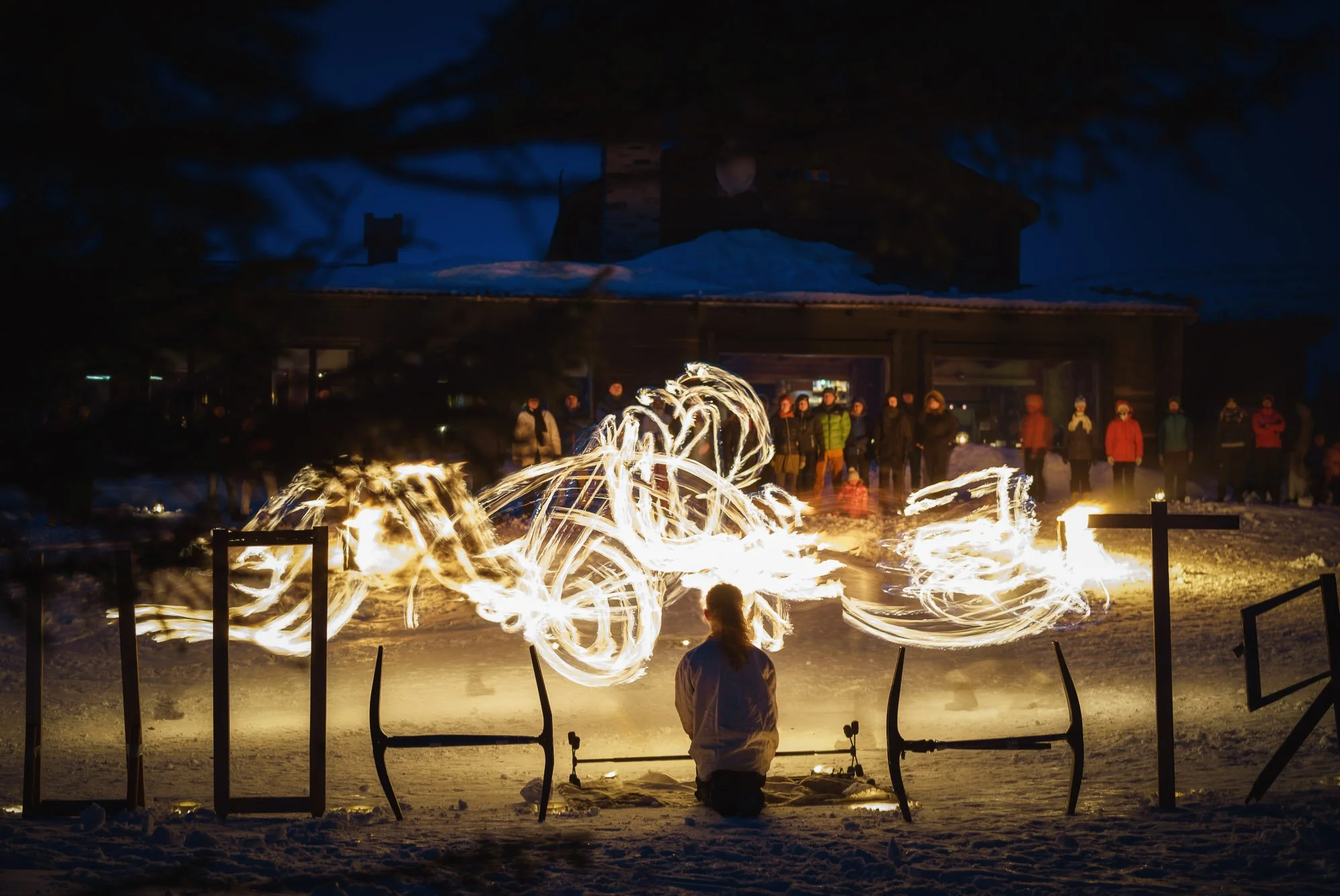 A person performing a fire dance during a winter night, with a crowd of onlookers in the background and snow on the ground.