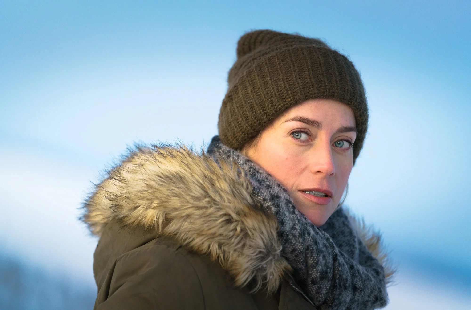 A woman in winter clothing, including a brown knit beanie, a scarf, and a coat with fur trim, outdoors in a cold environment with a blurry snowy landscape in the background.