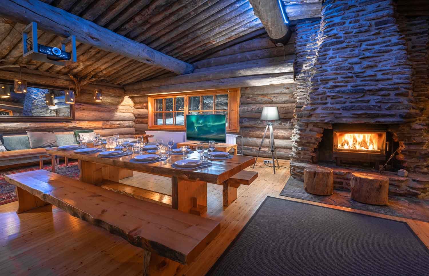 A cozy rustic dining room with a wooden table set for a meal, surrounded by benches and cushions, with a stone fireplace burning, logs and a window with curtains, and warm ambient lighting.