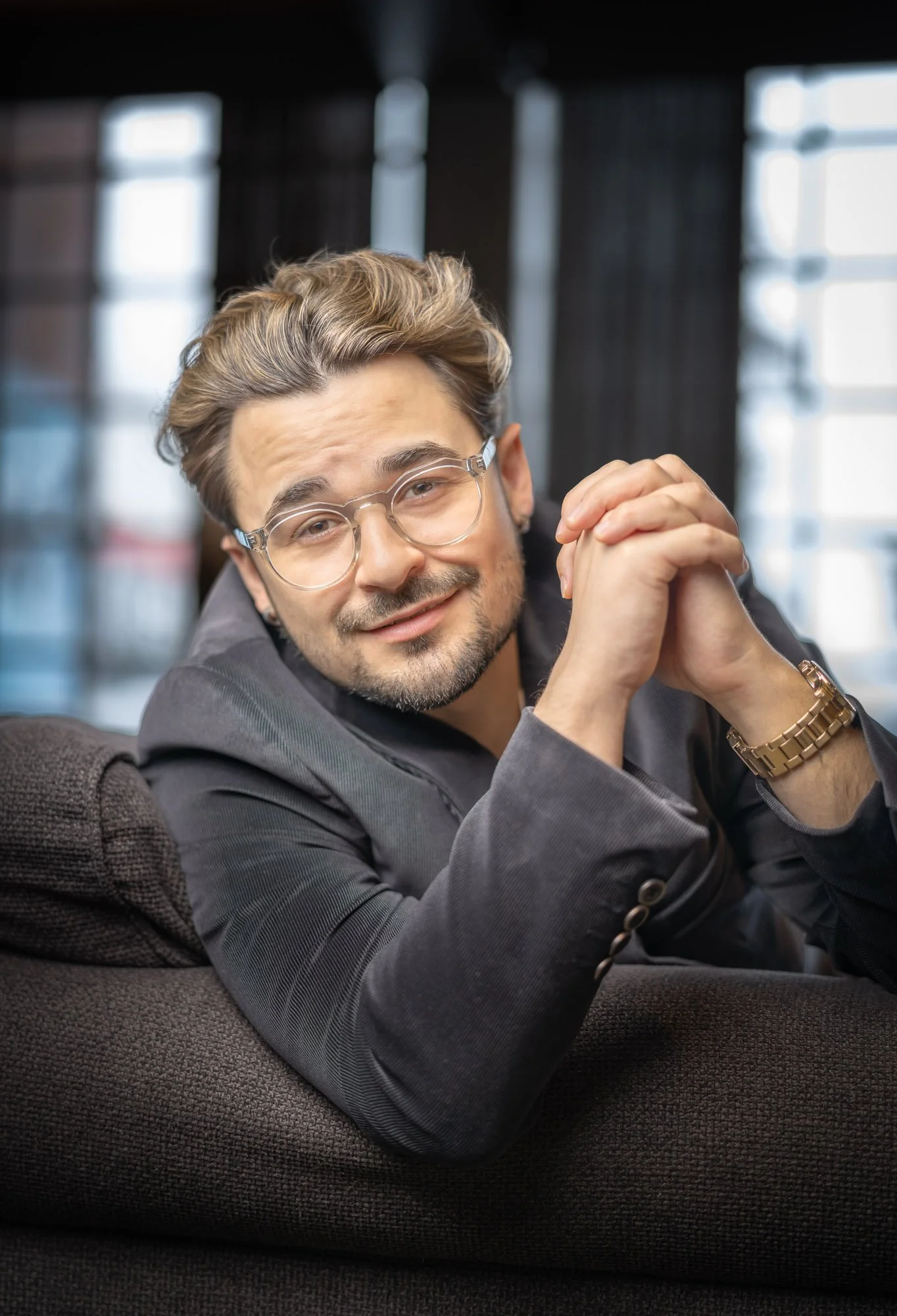 A young man with glasses and a beard, wearing a dark blazer, sitting on a couch, looking at the camera with a smile, in a modern indoor space with large windows in the background.