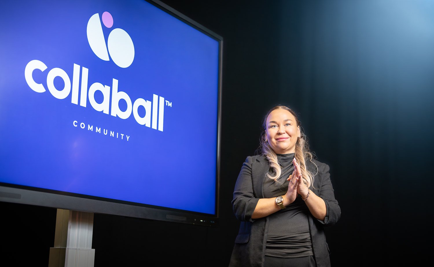 A woman stands beside a large display screen showing the Collaball Community logo, with her hands clasped together and smiling at the camera.