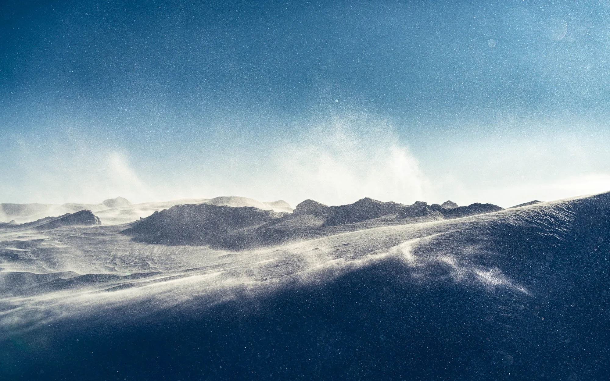 Snow-covered mountain landscape with windblown snow and a pale blue sky.