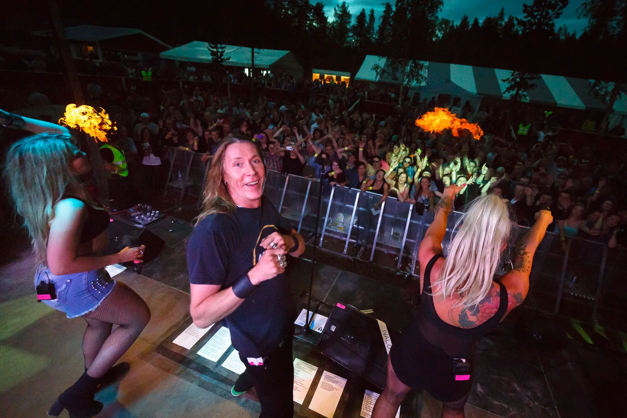 A female performer with blonde hair and tattoos on her back on stage, raising her arms, and a man with long hair, smiling and pointing, in front of a large crowd at a concert or music festival, with fire effects in the background.