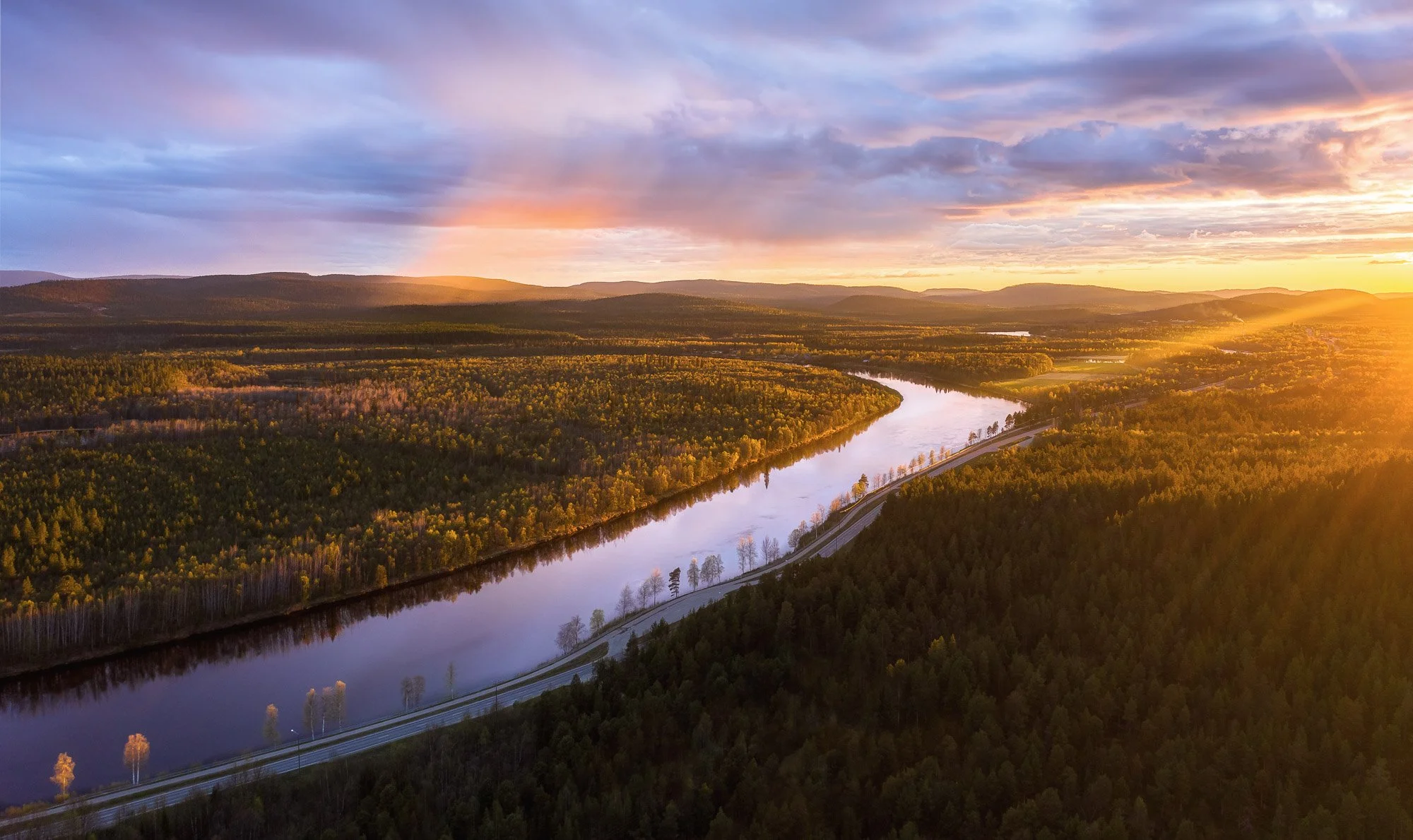 Aerial view of a winding river Ivalo flowing through a forested landscape at sunset with a road running alongside the river.