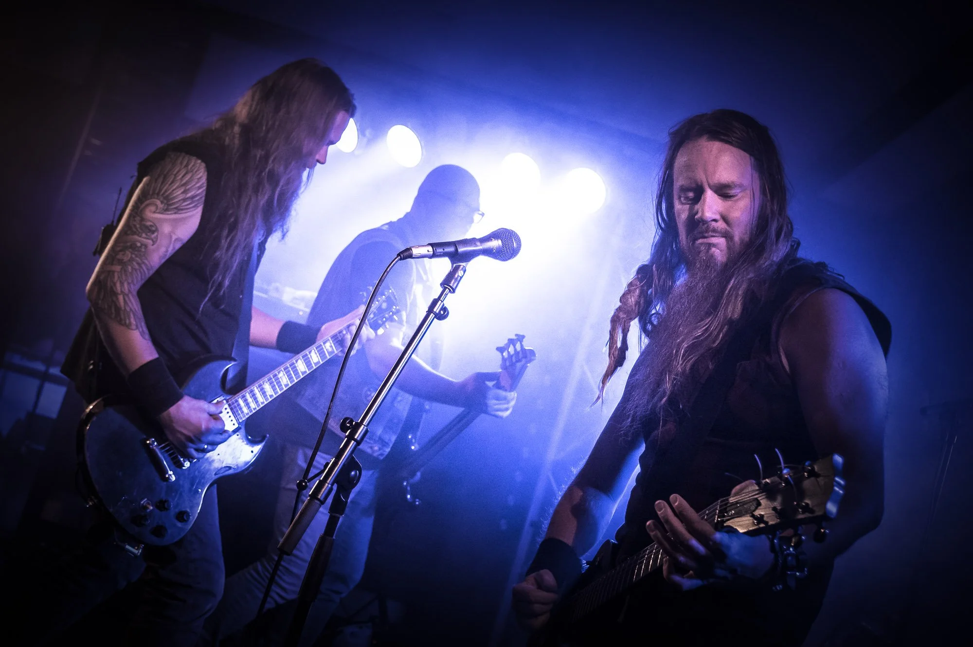 A band performs on stage with blue and purple lighting. Two guitarists are in the foreground, one with long hair and a tattooed arm, and the other with long hair and a beard. In the background, a person with glasses plays drums.