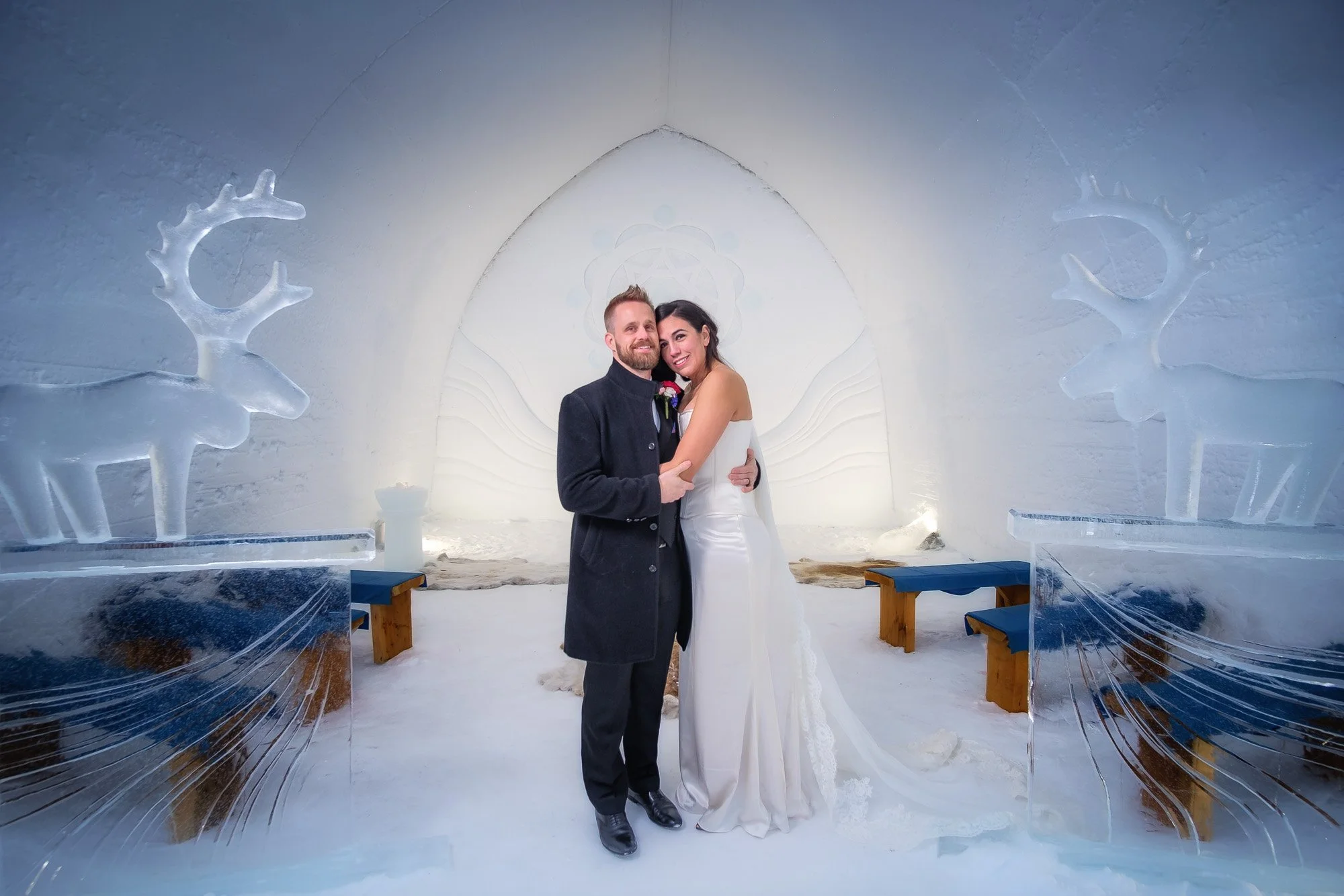 A couple dressed in wedding attire standing close together inside an ice chapel with ice sculptures of reindeer on either side, with white snow and ice decor surrounding them.