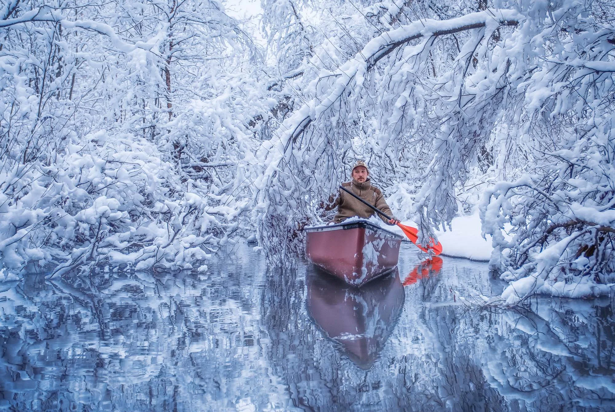 A person paddling a boat through a snow-covered river surrounded by snow-laden trees.