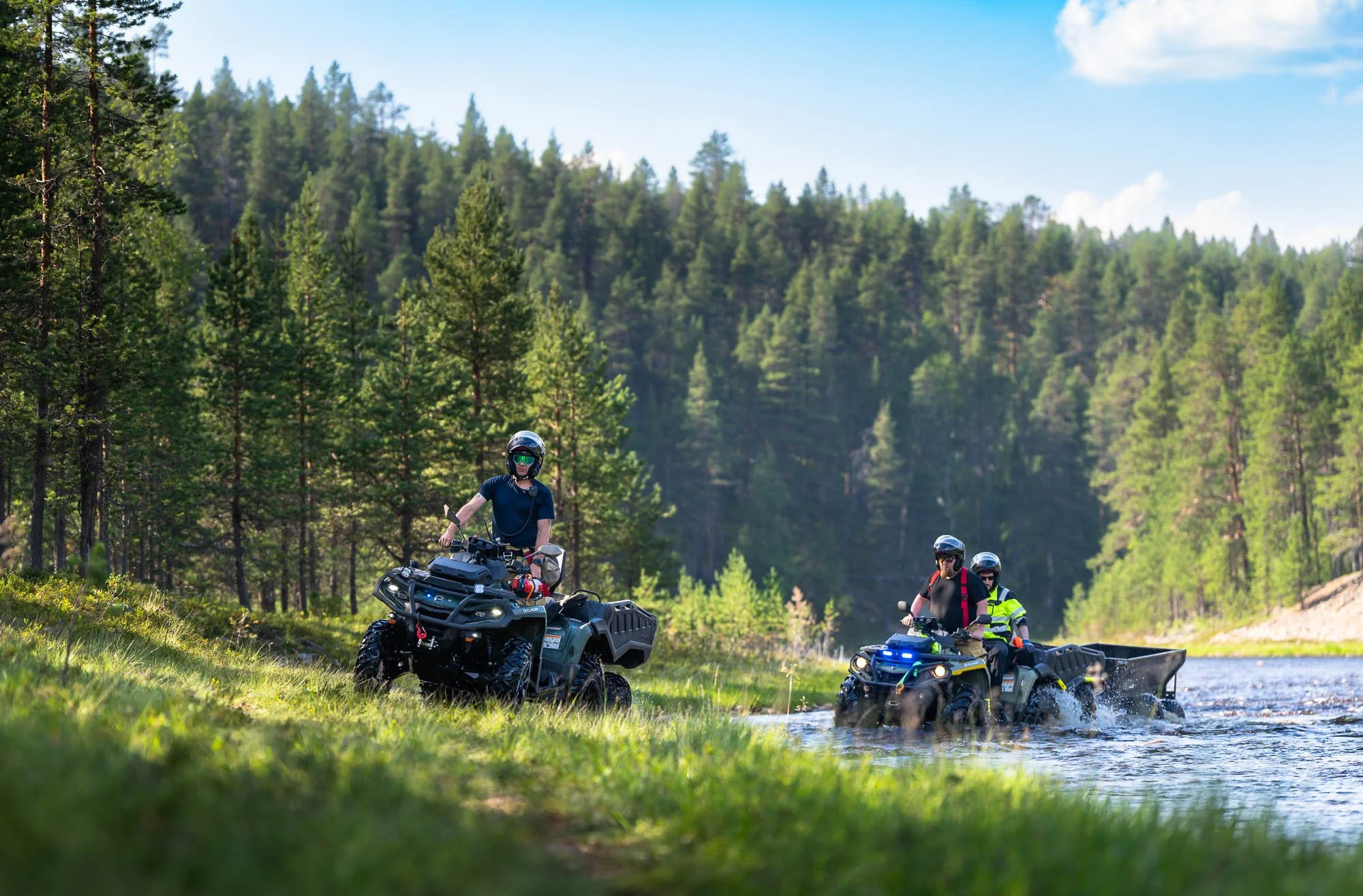 Three people riding all-terrain vehicles near a river in a lush, green forest on a sunny day.
