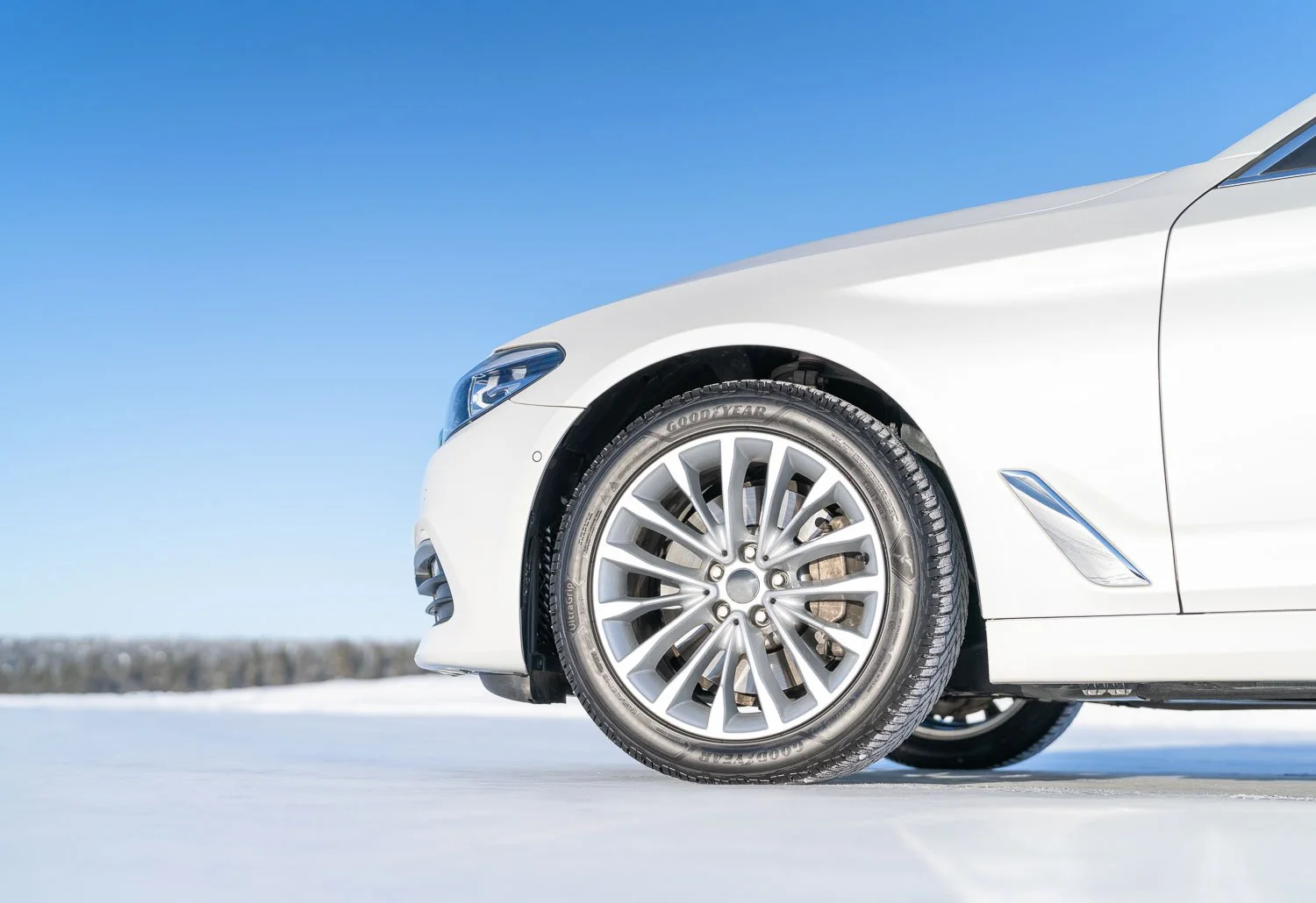 Close-up of the front right side of a white car with alloy wheel and tire, parked on snow under a clear blue sky.