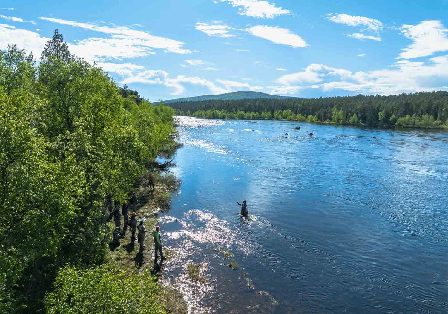 People gather along the riverbank to watch a person fly fishing in the water, with lush green trees on the left, blue sky with scattered clouds, and a distant mountain range in the background.