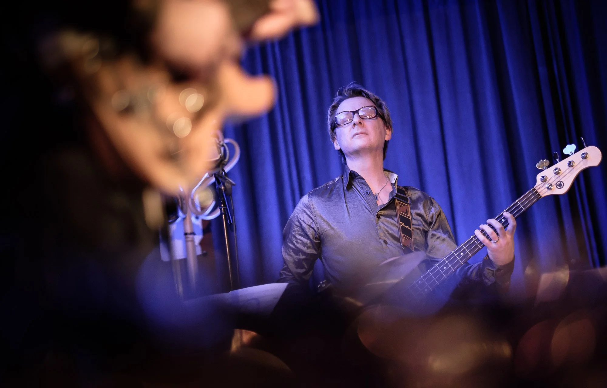 Musician with glasses playing a bass guitar on stage with a blue curtain background.
