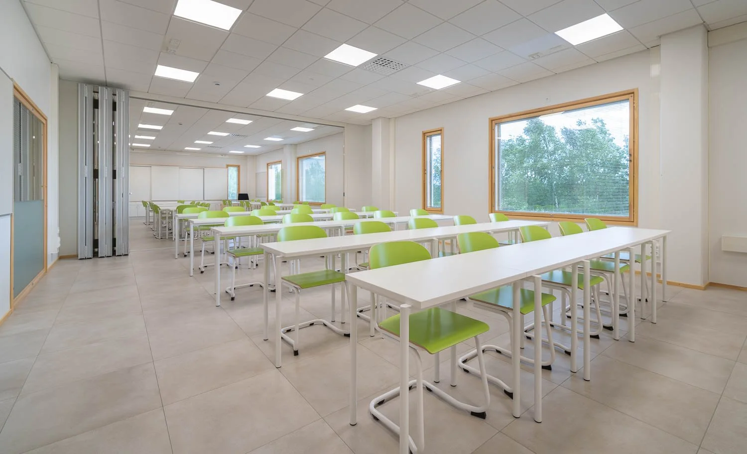 Empty classroom with white tables and green chairs, large windows, and a white ceiling with fluorescent lights.