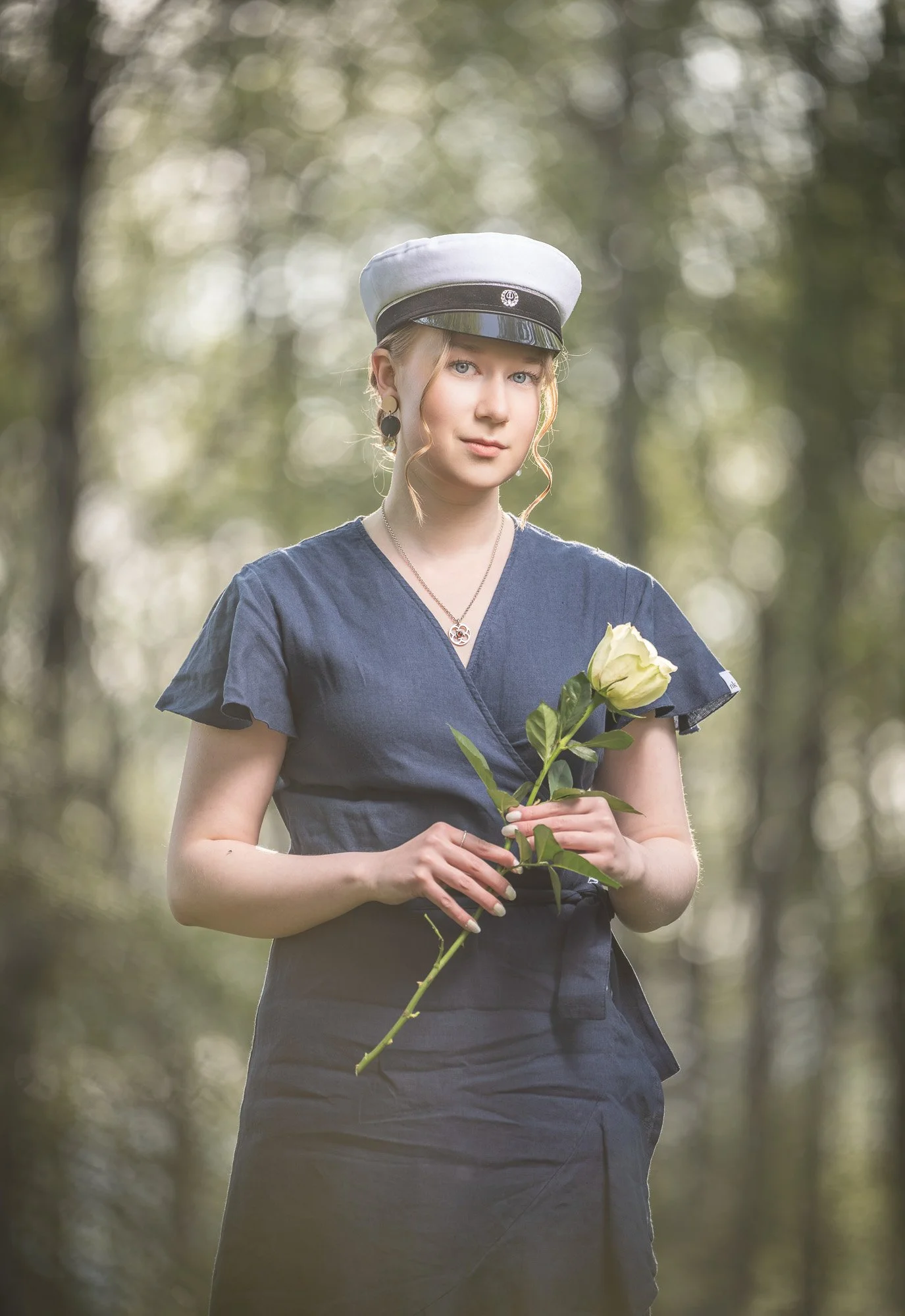 A young woman with blonde hair, wearing a white and black hat, a navy blue dress, and black earrings, stands outdoors in a wooded area holding a white rose.