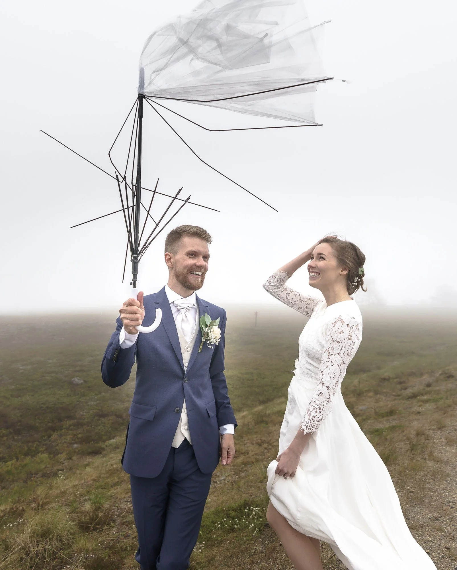A smiling couple dressed in wedding attire standing outside in a foggy, grassy field. The man is holding an open black umbrella, and the woman is touching her hair while smiling at him.