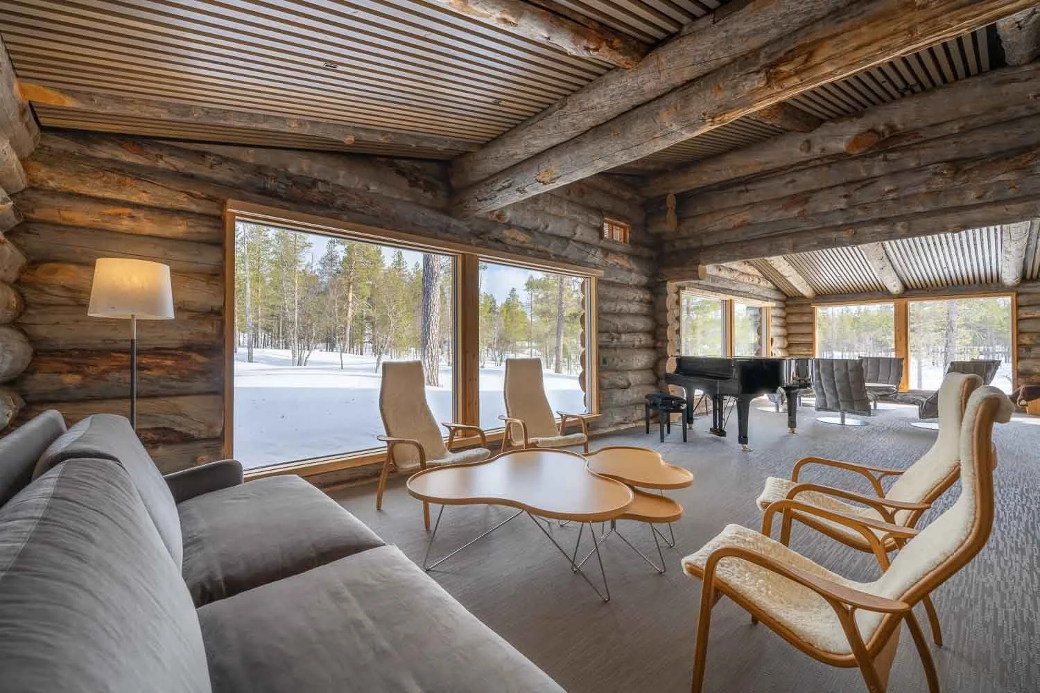 Interior of a rustic log cabin living room with large windows showing snow-covered landscape and trees outdoors. Contains a gray sofa, armchairs, a black grand piano, and a modern wooden coffee table. A standing lamp is beside the sofa.