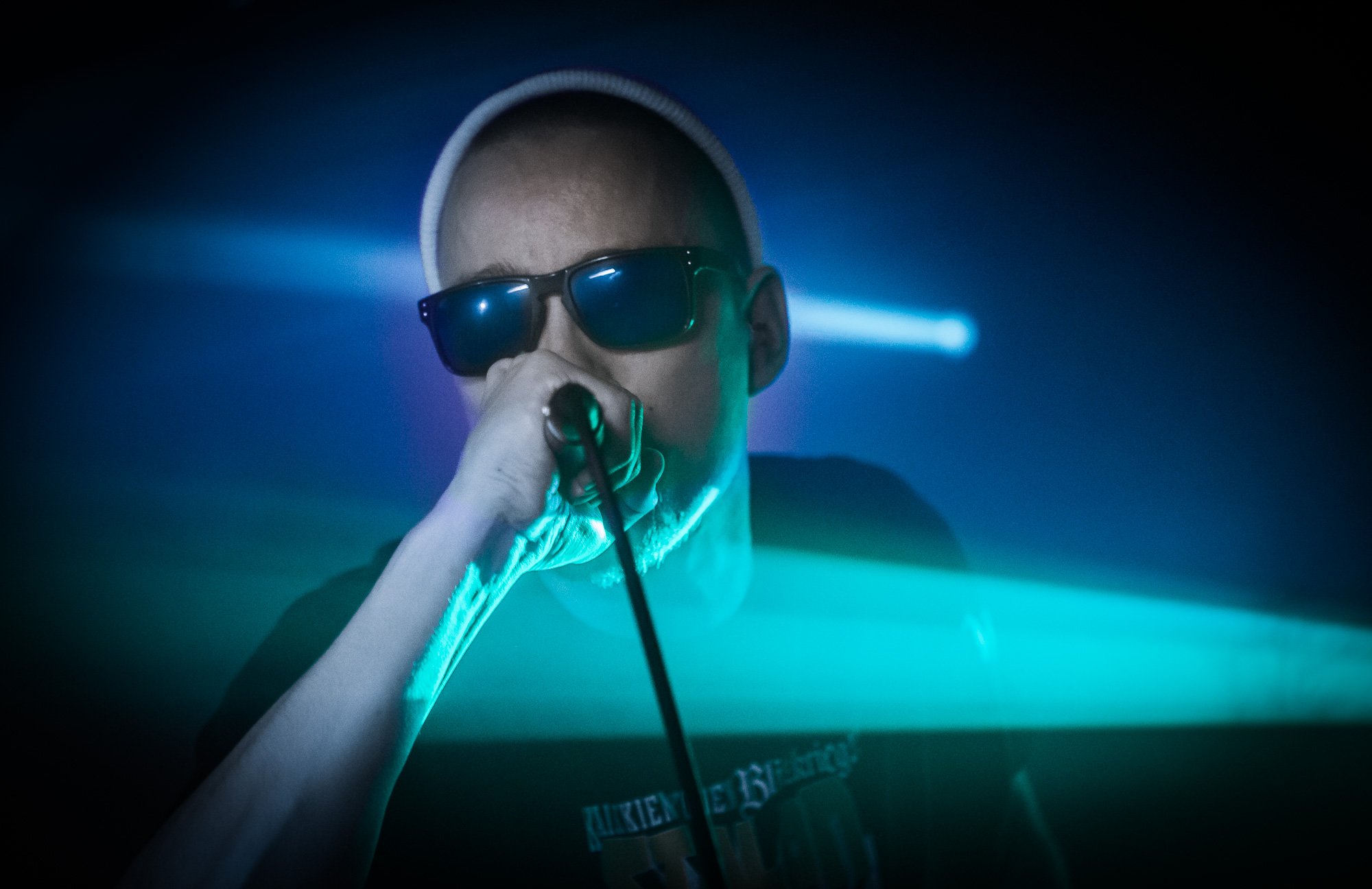 A person wearing sunglasses and a white cap, holding a microphone, illuminated by colorful stage lights in a dark setting.